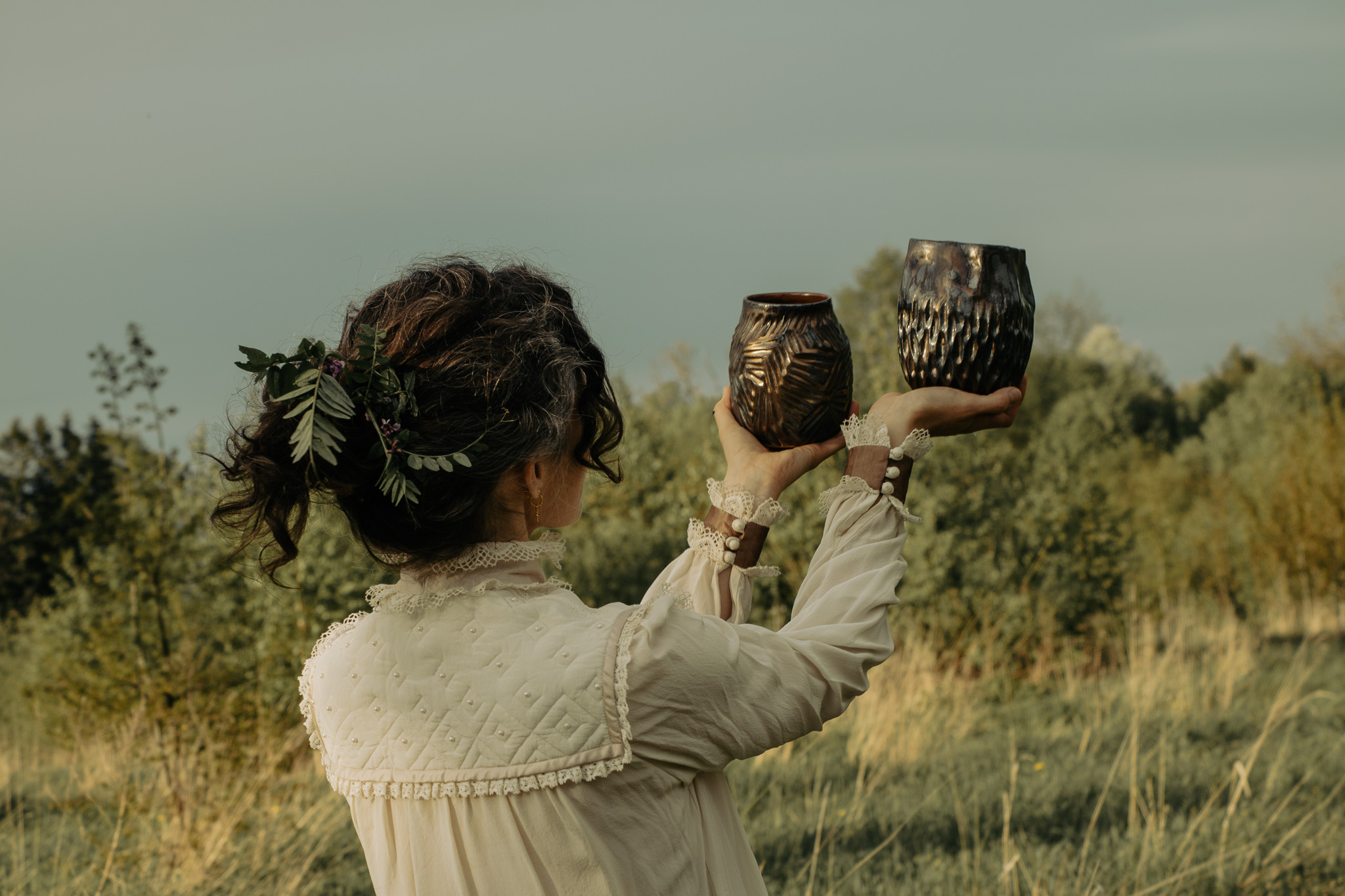 Victoria holding two dark handmade ceramic vessels in a summer field